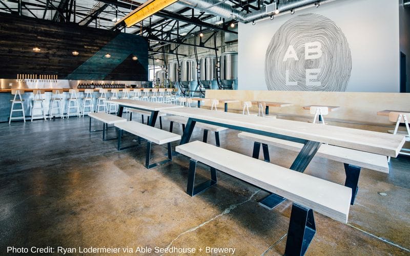 Able Seedhouse and Brewery is one of the best Minneapolis breweries, located in the Northeast neighborhood. Concrete floors with long wood tables on dark metal frames. In the distance a metal bar lined with chairs and a white sign with the Able logo.