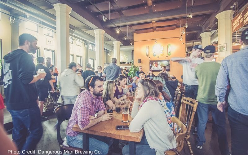 Dangerous Man Brewing Co is one of the best Minneapolis breweries, located in the Northeast neighborhood. A small taproom filled with people at wooden community tables with mismatched chairs. White columns separate a seating area that lines the windows.