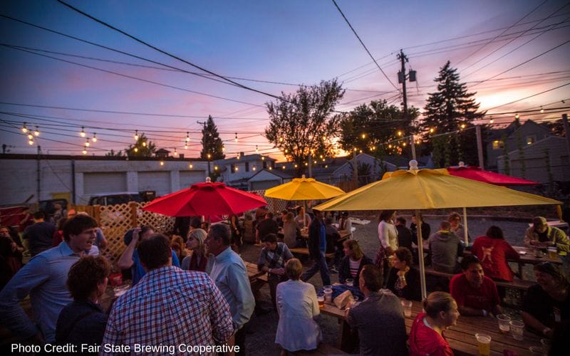 Fair State Brewing Cooperative is one of the best Minneapolis breweries, located in the Northeast neighborhood on Central Ave. Pink, blue and purple sunset sky above the red and yellow umbrellas and wooden picnic tables in Fair State Cooperative's outdoor patio space.