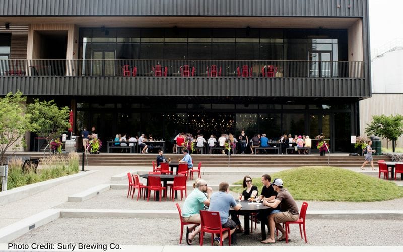Surly Brewing Co. is one of the best Minneapolis breweries, located close to the University of Minnesota campus. Surly's taproom as viewed from the outside is a two story covered patio along  a black metal building. The tables and chairs are bright red. In front of the building is a grass and gravel outdoor space with additional tables.