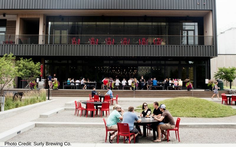 Surly Brewing Co. is one of the best Minneapolis breweries, located close to the University of Minnesota campus. Surly's taproom as viewed from the outside is a two story covered patio along a black metal building. The tables and chairs are bright red. In front of the building is a grass and gravel outdoor space with additional tables.