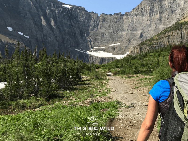 A woman in the lower right hand corner is hiking with a backpack on. The trail extends off into the distance to the base of the mountains that are partially covered in snow.