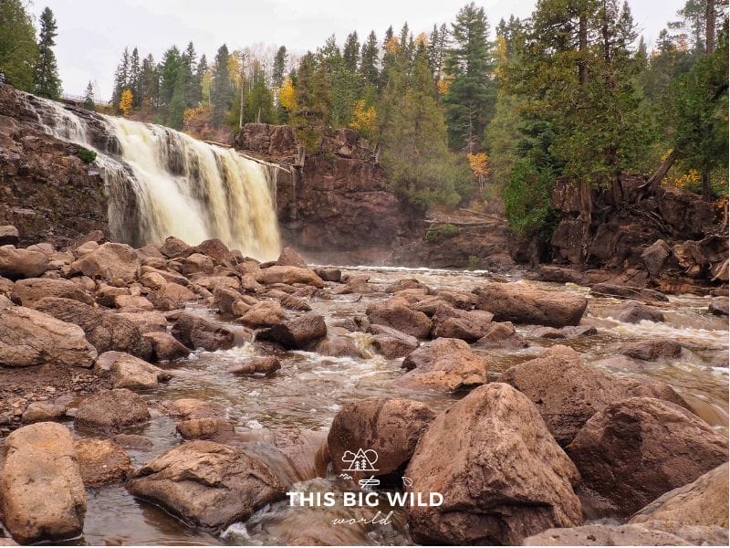 To the left in the distance is a tall waterfall lined on either side with yellow and green pine trees. In the foreground, the water flows over large rocks at Gooseberry Falls State Park along Minnesota's North Shore of Lake Superior.