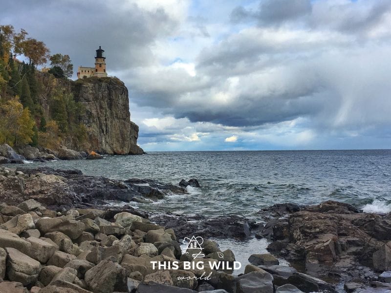 From the left is a granite rock formation with a peach lighthouse, overlooking the dark water of Lake Superior. In the foreground is a rocky beach and in the distance to the right is a peek at a blue sky through the dark stormy clouds at Split Rock Lighthouse State Park along Lake Superior in Minnesota.