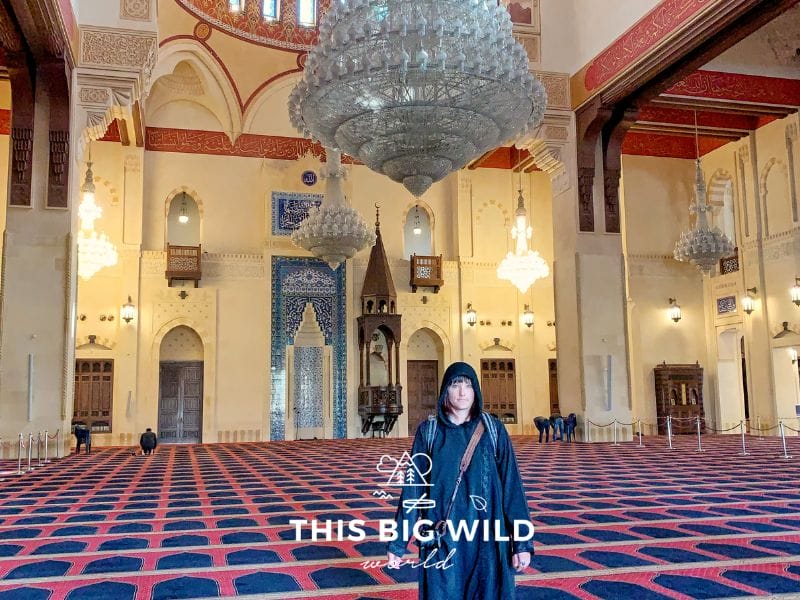 Me wearing a black cloak and head covering inside Mohammed Al Amin mosque in Beirut. Extravagant crystal light fixtures hang from the ceiling over the prayer space.
