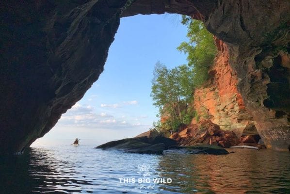 A kayaker paddles through a red sandstone arch of a sea cave along the shore of Lake Superior.