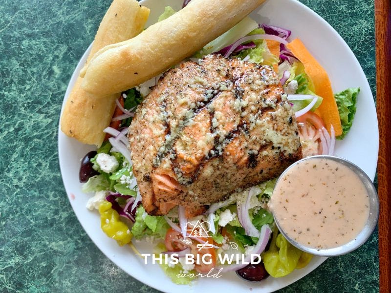 A plate with a greek salad with grilled salmon on top and a side of two breadsticks and a vinaigrette dressing from Apollo Restaurant in Seward.