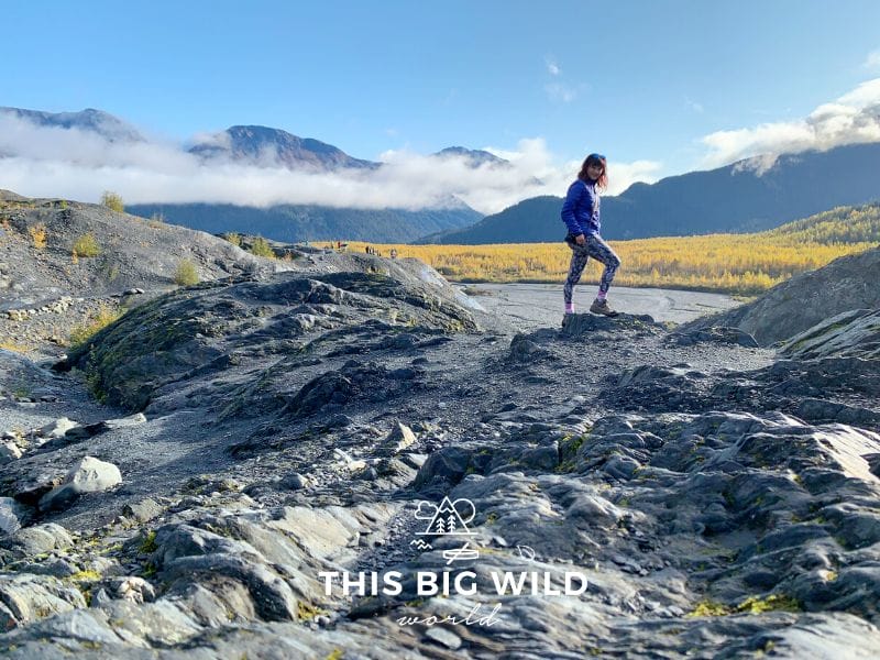 Me hiking on a rocky surface with yellow fir trees and mountains with low cloud cover in the background.