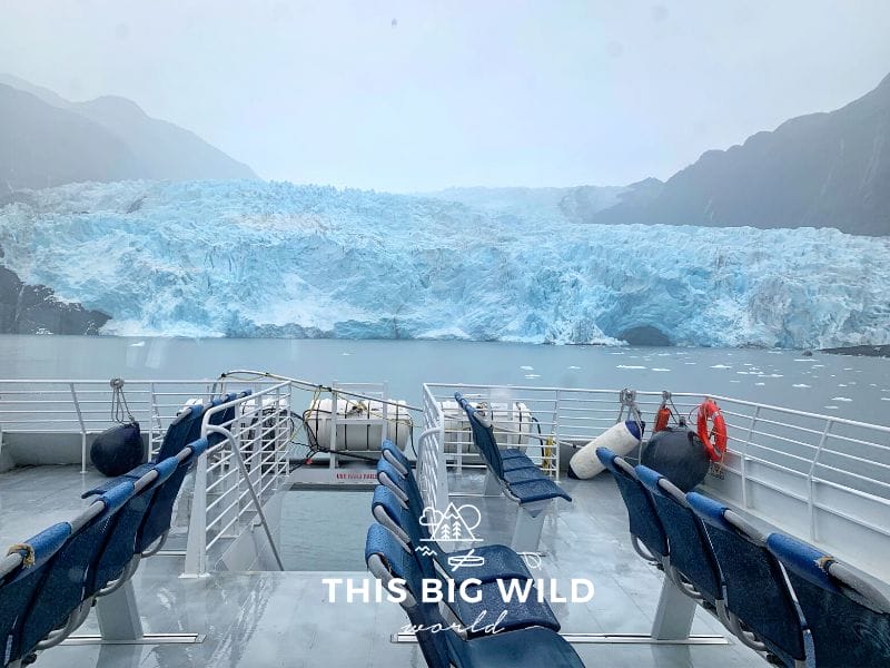 Ailik Glacier is bright blue, even on a rainy gray day, as viewed from the back of a boat with dark mountains on either side.