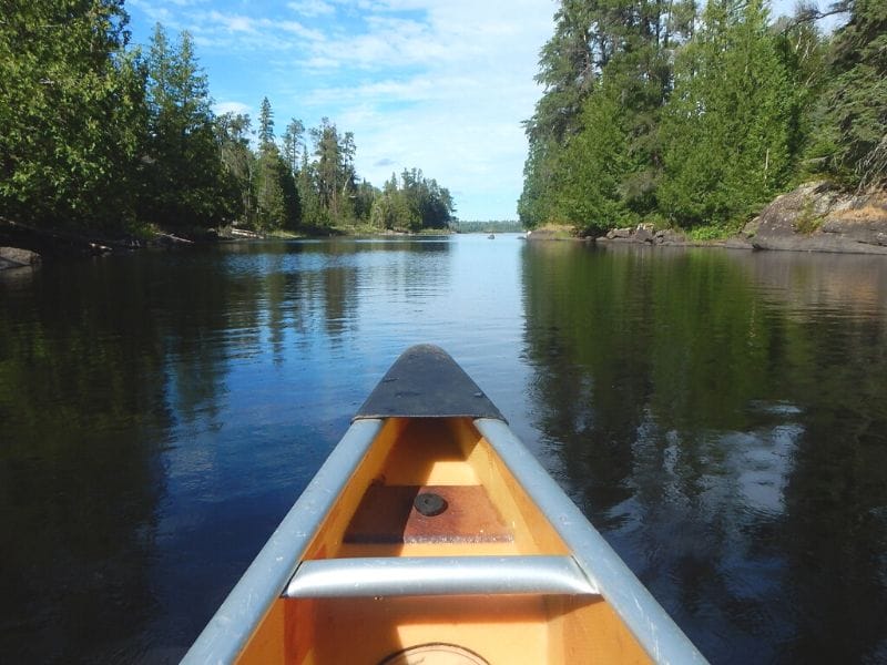 View from inside a canoe on the water in the Boundary Waters Canoe Area in northern Minnesota. Trees line both sides of the water on a sunny day. This is the number one item on any outdoor adventurers' Minnesota bucket list!