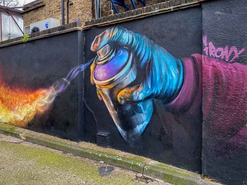 A dark blackish wall painted with vibrant colors in Camden, London. On the right a bright pink and purple arm is holding a large spray can with a blue gloved hand. The spray can is spraying bright red-orange resembling flames. Photo credit: Wayfaring Views.
