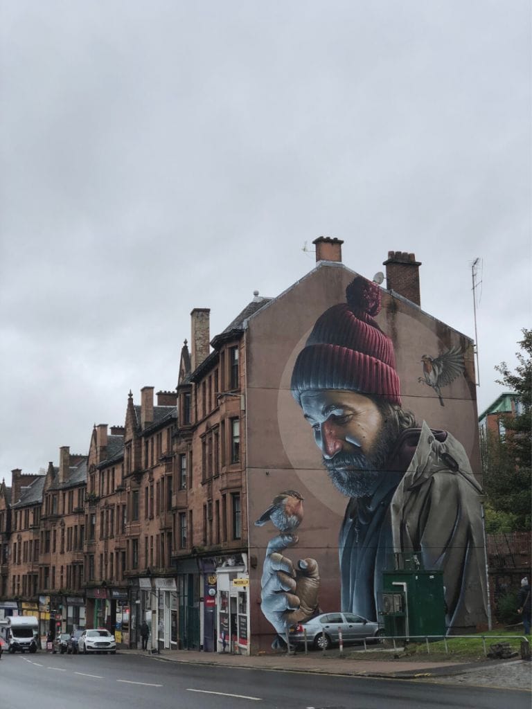 A row of flats in Glasgow is lined with shops on the ground floor on a gray cloudy day. On the end of the building is a large painting of a man in winter hat holding a small bird in his hand. This is one of many stops on a Street Art tour in Glasgow with Airbnb Experiences. Photo credit: Laura No Esta