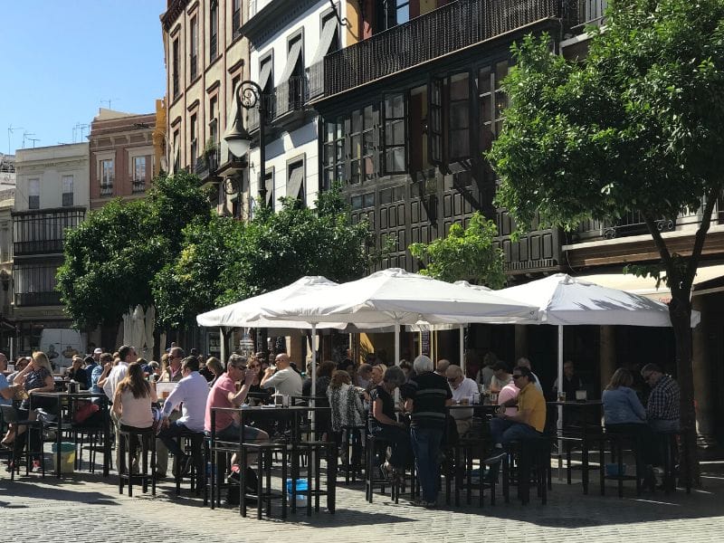 Lining the sidewalk in front of a row of old colorful buildings in Seville, are tables filled with locals enjoying tapas and drinks on a sunny day. Photo credit: Where Angie Wanders