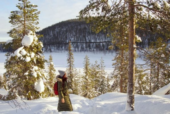 How to choose between Levi and Rovaniemi when visiting Lapland in winter! Image of me hiking in deep snow in a forest overlooking a frozen lake with sun shining on me.