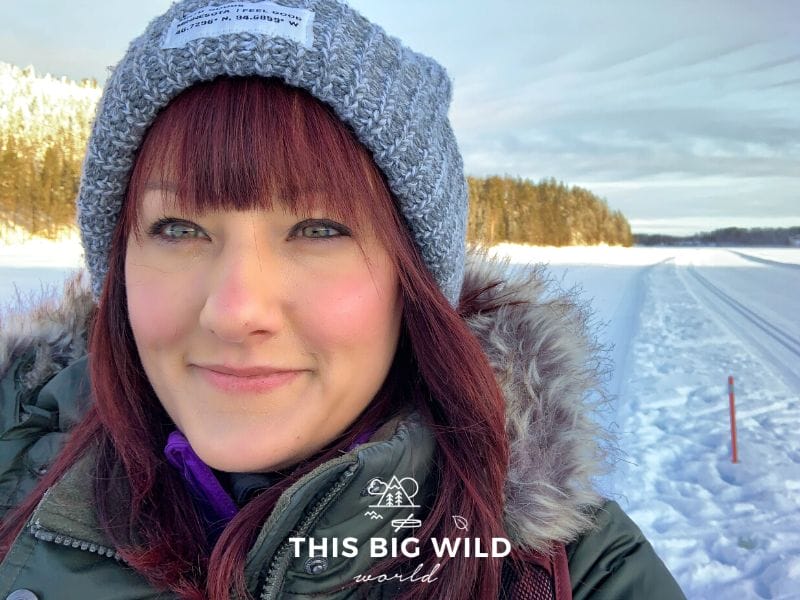 Close up shot of me, a woman with bright red hair, a gray winter hat and green puffy coat with a fur around the hood. Behind me is a frozen lake with sun hitting a the snow covered fell lined with pine and fir trees.