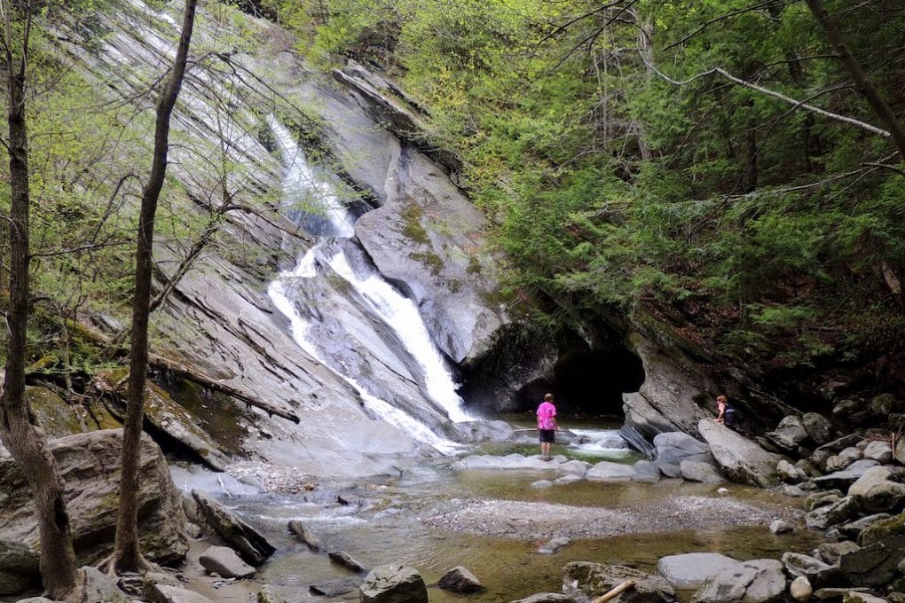 A dramatic waterfall flows of larger than life stones into shallow pools at the bottom. Lush green trees are on either side of the waterfall and a woman in pink is standing on a rock in the water enjoying the view.