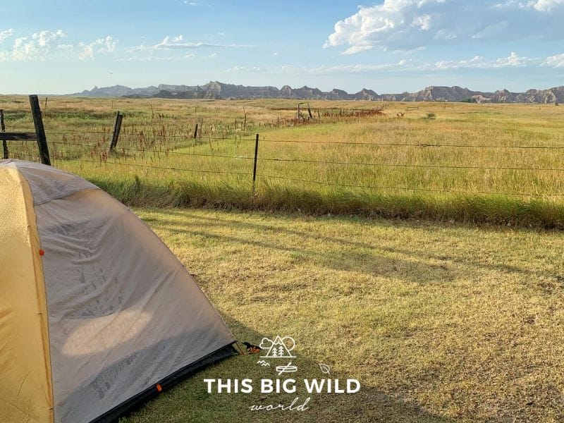 In the left foreground is a gray and yellow tent setup on grass. Behind it is a wire fence with taller green grass on the other side. In the distance across the frame is dark reddish outline of the rock formations in Badlands National Park. White clouds scattered across a blue sky are overhead. The sun is coming into the frame from the left hand side.