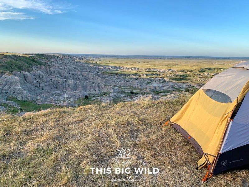On the right hand side, a gray and yellow tent is setup on dry grass at the edge of a cliff overlooking the sweeping Badlands rock formations below. In the distance are RVs and other tents along the cliff. Below is green grass with bison roaming. The sun is coming into the frame from the left hand side.