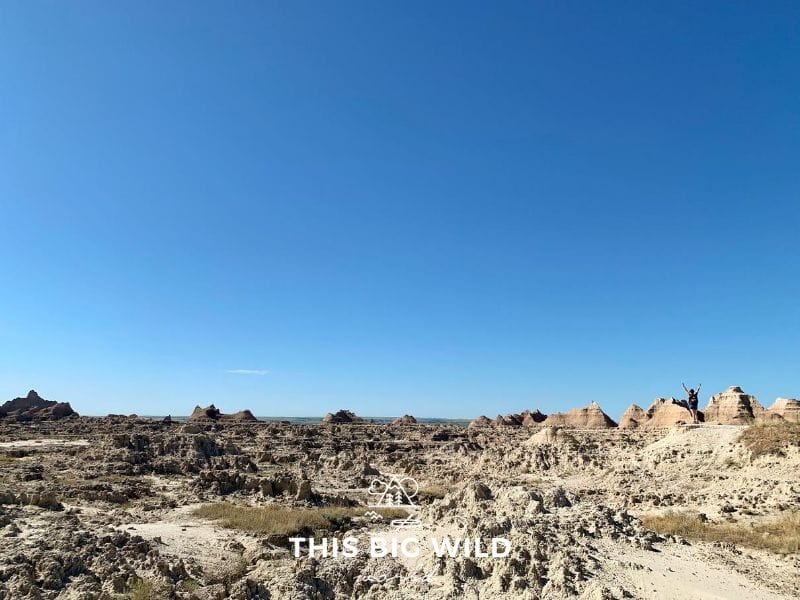 The lower third of the image shows jagged clay rock formations all the way to the horizon. The upper portion of the image is clear blue sky. On the right hand side, I am standing, looking very small standing on one of the rock formations with my hands raised over my head.