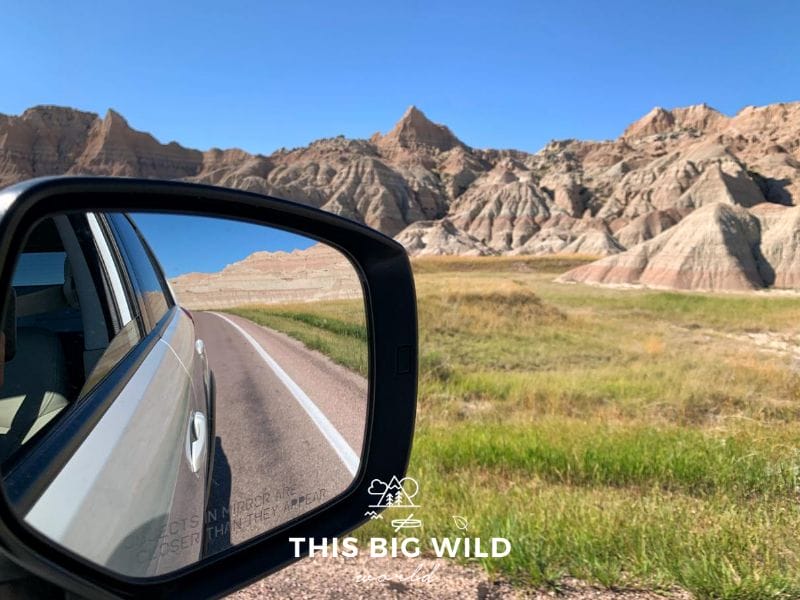 The left hand side of the photo is a closeup of a rear view mirror on a white vehicle, showing the open road and jagged rock formations in the reflection. The right hand side show the landscape on the road ahead, with colorful rock formations, green grass and a bright blue sky.