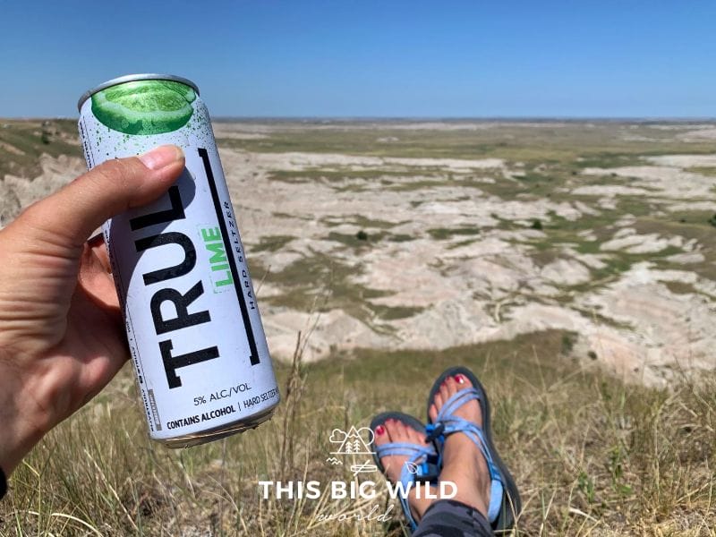 On the left side of the frame, my hand is holding a Truly lime hard seltzer can. At the bottom of the image you can see my feet stretched out in the grass with blue Chaco sandals. In the distance is a valley below with rocks formations, grassland and grazing buffalo.