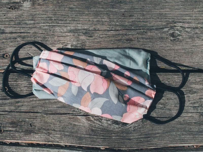 Two face masks rest on top of each on wooden planks. The masks have pink, green and gray floral design and black ear straps. The best face mask for hikers has 3 layers of fabric but is still breathable and comfortable.