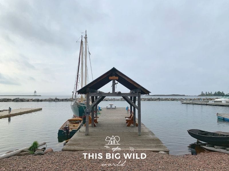 In the center, a rocky shoreline gives way to a wooden pier with a small shelter built over it. A wooden bench is on either side of the pier. And on the left side are two boats, one motorize and one is a sailboat.In the distance is a rocky breakfast and a small lighthouse.