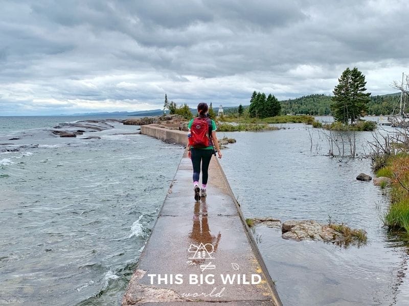 In the center of the image a concrete narrow walkway stretches out into the water, with water on both sides. There are small waves in the dark blue water and cloudy sky overhead. In the distance is small white lighthouse and green trees, jagged rocks peek out from the water. In the center, a female is walking with black pants, a green shirt and bright red backpack on.