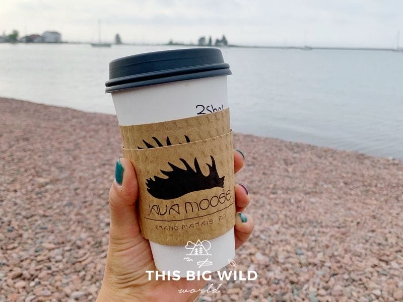 A woman's hand with teal nail polish holds a disposable cup of Java Moose coffee with a rocky shore and Lake Superior in the background.