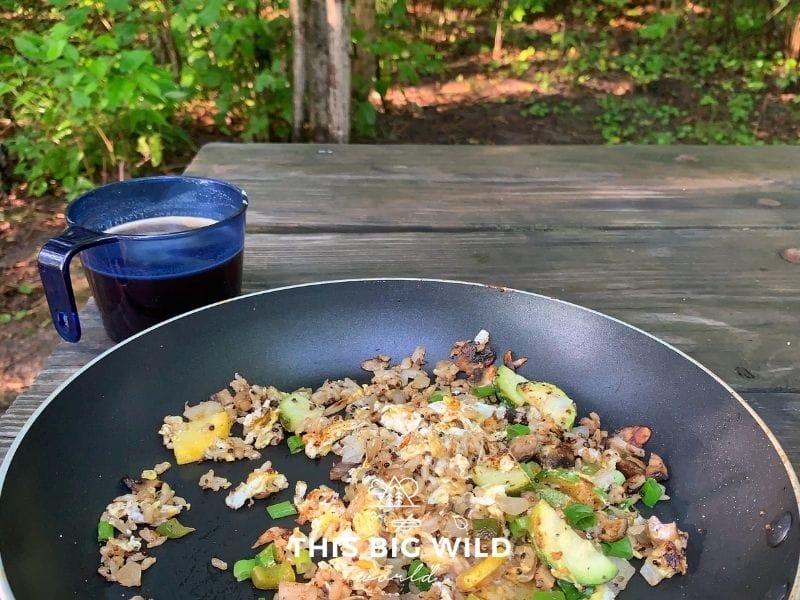 A skillet with eggs, quinoa, and vegetables sits on a wooden picnic table with a blue plastic mug filled with coffee. Green trees and forest are in the background.