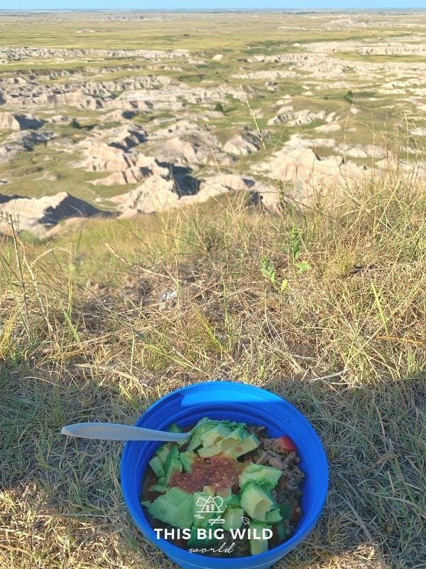 A blue Sea to Summit collapsible camping bowl is filled with a burrito bowl, topped with fresh avocado and salsa. The bowl is sitting in brown grass overlooking the Badlands.
