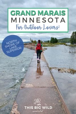Text: Grand Marais Minnesota for Outdoor Lovers!
Subtext: North shore of Lake Superior
Background Image: A woman walks on a narrow concrete path with water from Lake Superior on either side. In the distance is a green forest a small white light house and a cloudy dark blue sky overhead.