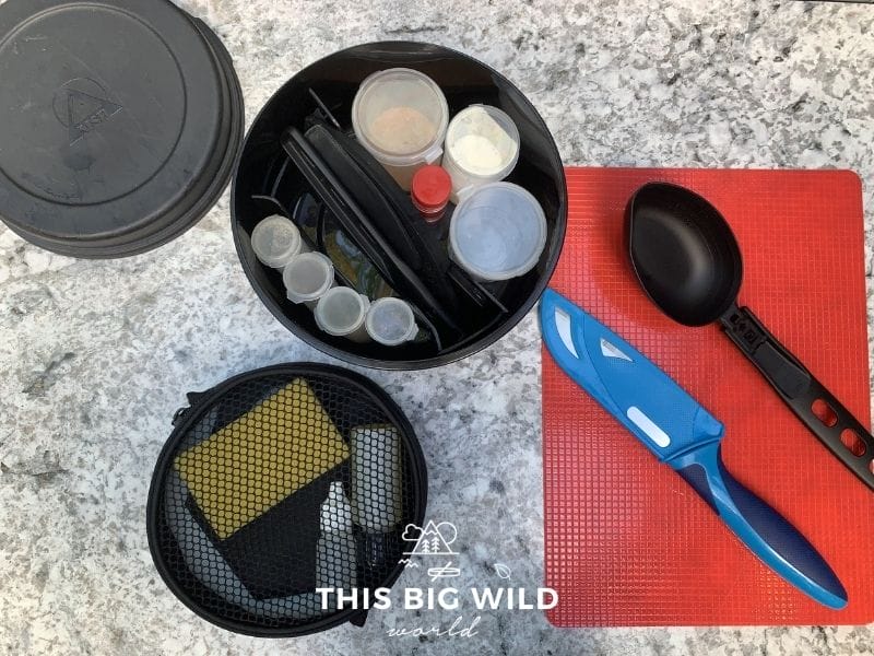 An overhead photo of an MSR kitchen kit. In the top left is the black plastic lid for the kit. In the center is the kit with 4 small clear plastic bottles to hold spices, 2 foldable cooking utensils, and 3 larger clear plastic bottles for more spices all in a round black container. To the right is a cutting board, foldable cooking spoon, and cutting knife with removable cover. In the bottom left is a black round mesh container holding a dish washing pad, dropper for dish soap and cutting board.