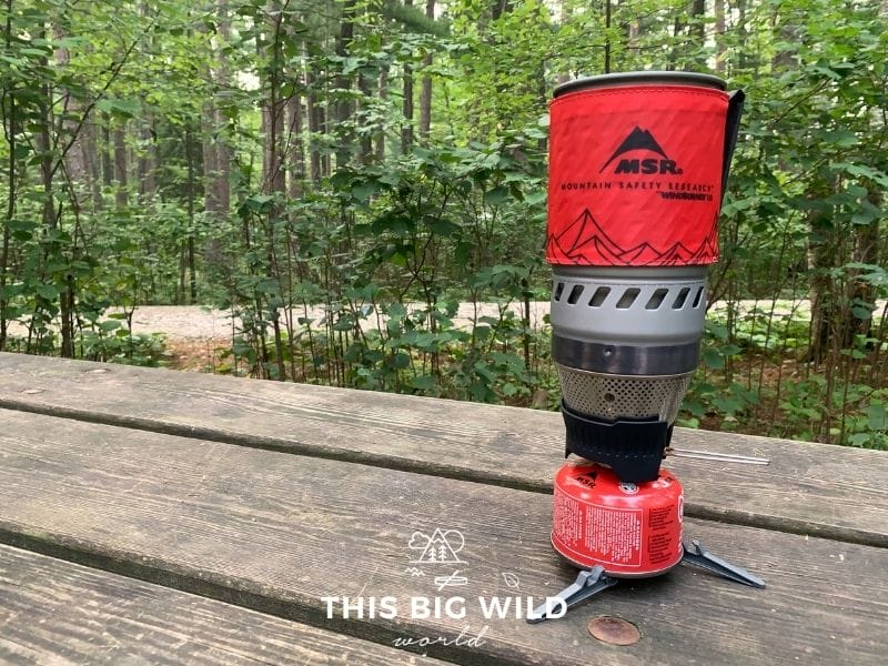 The MSR Windburner backpacking stove has a small red fuel canister, black and silver burner and an insulated red container that sits on top to boil water in. The stove is sitting on a wooden picnic table with green trees behind it.