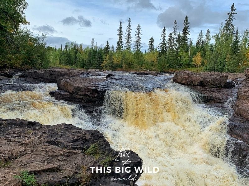 A wide waterfall flows through dark brownish red rock with smaller waterfalls overflowing on either side. The water is slightly brown and flowing quickly. On the horizon is a line of green pine trees with a bright blue sky overhead.