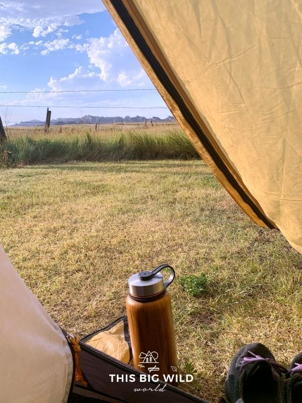View from inside of a tent looking out towards a mountain range in the distance and blue skies overhead. Next to the tent is a wooden finished water bottle and sneakers sitting on the grass.