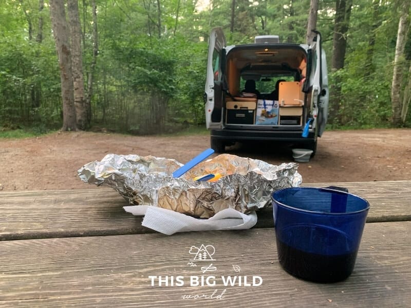 A foil packet dinner and blue plastic cup sits on a wooden picnic table in the foreground. In the background is the rear of the campervan open with lights on showing a small sink and refrigerator. 