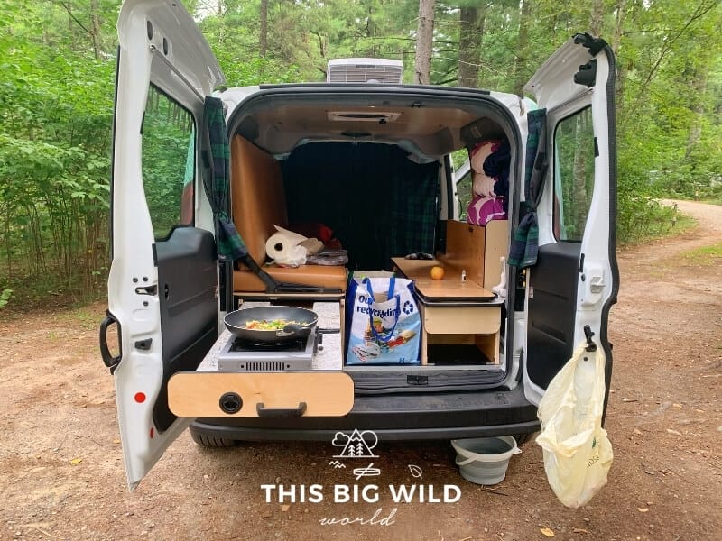 The rear doors of a campervan are open showing the countertop pulled out. Food is cooking in a skillet on a burner on the countertop. The bed is setup as a bench with cooking supplies sitting on it. The van is parked at a campsite in a forest.