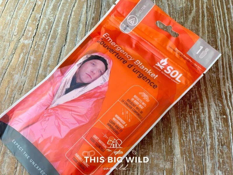A small bright orange plastic pouch is laying on a wooden surface. It's labeled an Emergency Blanket for one person.