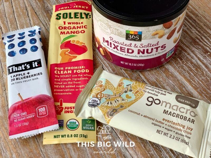An upclose shot of various food items laying on a wooden surface, including a can of roasted and salted mixed nuts, an everlasting joy GoMacro bar, a mango fruit jerky by Solely, and an apple blueberry That's It bar.