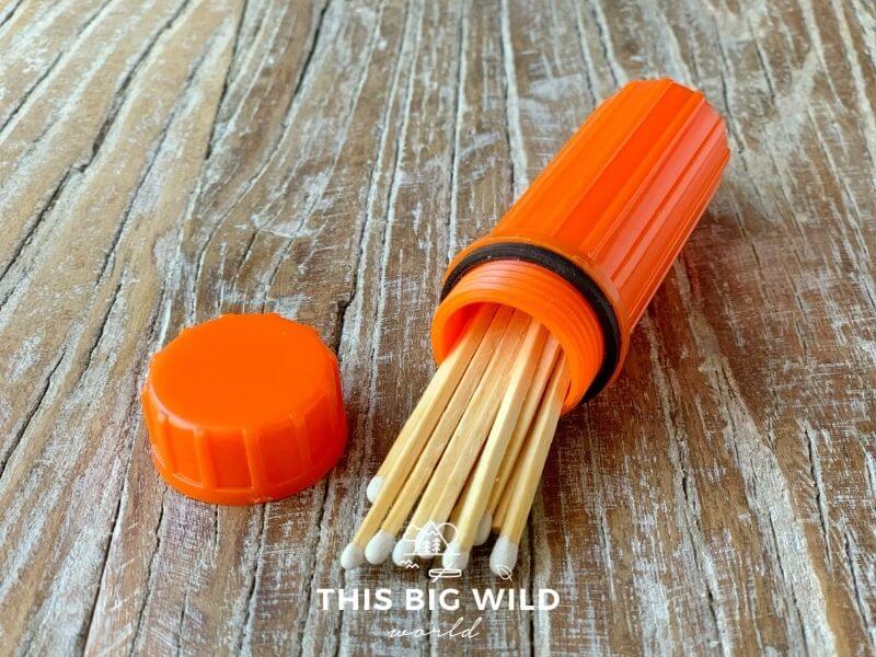 A bright orange container is laying on its side on a wooden surface. Wooden matches are coming out of the end of the container and an orange screw on cap is sitting next to it.