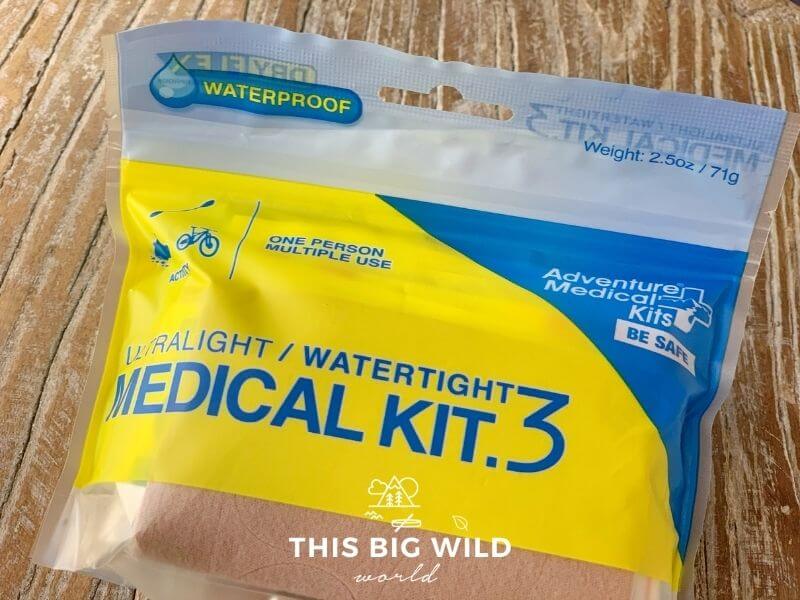 An close up shot of a small First Aid kit in a plastic waterproof pouch lying on a wooden surface. The package is bright yellow with some blue on the top. It's labeled the Ultralight/ watertight medical kit.3 by Adventure Medical Kits.