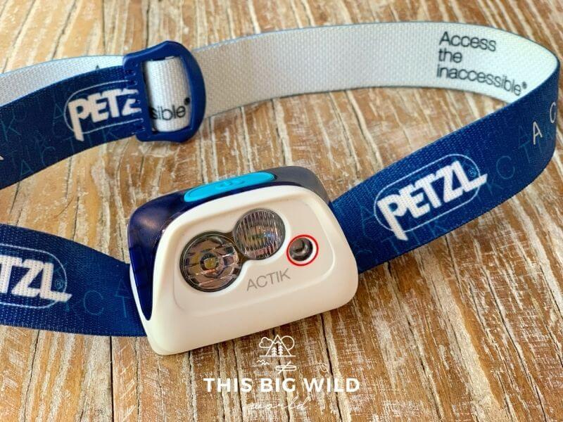 An up close shot of a blue and white headlamp laying on a wooden surface. The band is blue on the outside with the Petzl logo and white on the inside. The lamp itself is white with a blue button and three different bulbs.