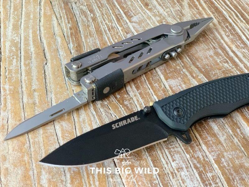 Two knives are laying diagonally on a wooden surface. At the top is a silver multi-tool with the knife extended on one end and the pliers on the other end. At the bottom is a black single foldout knife by Schrade.