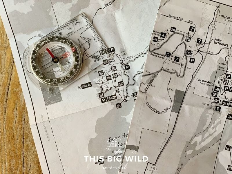 Two paper hiking maps are laying on top of each other on a wooden surface with a clear plastic compass lying on top of them both.