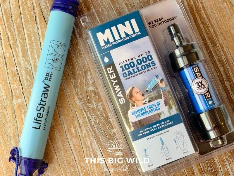 On the left is a LifeStraw which is a light blue tub with a blue cap on either end. On the right is a Sawyer Mini water filtration system in its packaging. Both are laying on a wooden surface.