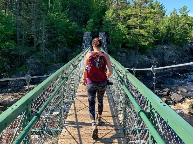 A wood bridge with green metal rails extends from the center of the frame straight across into the distance. At the far end is a green forest and below is a rocky stream. A woman (me) is walking across the bridge with a red backpack on carrying a water bottle, handheld GPS and safety whistle attached to it.