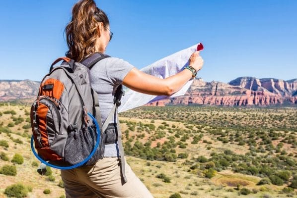 A woman is hiking in desert area with mountains in the distance. Her back is turned to the camera. She's wearing a backpack and looking at a map.