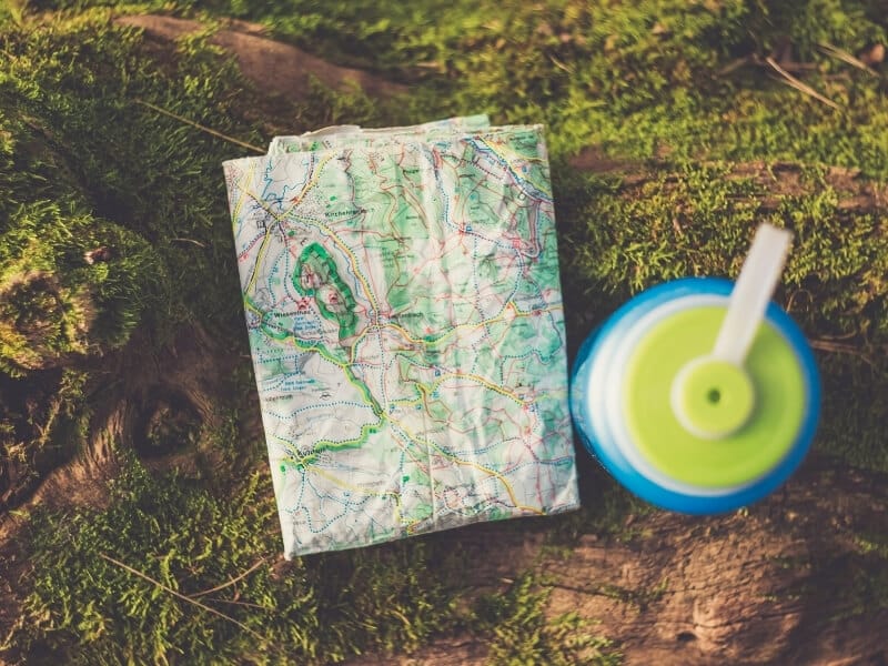 A paper trail map is folded and faded laying on a green mossy surface next to a blue and lime green water bottle.