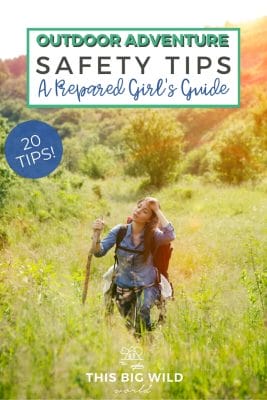 Text: Outdoor Adventure Safety Tips - A Prepared Girl's Guide. 20 Tips!
Image: A woman walks through tall grass wearing a backpack and carrying a walking stick. Behind her is a large hill and in the sun setting in the top right of the frame.